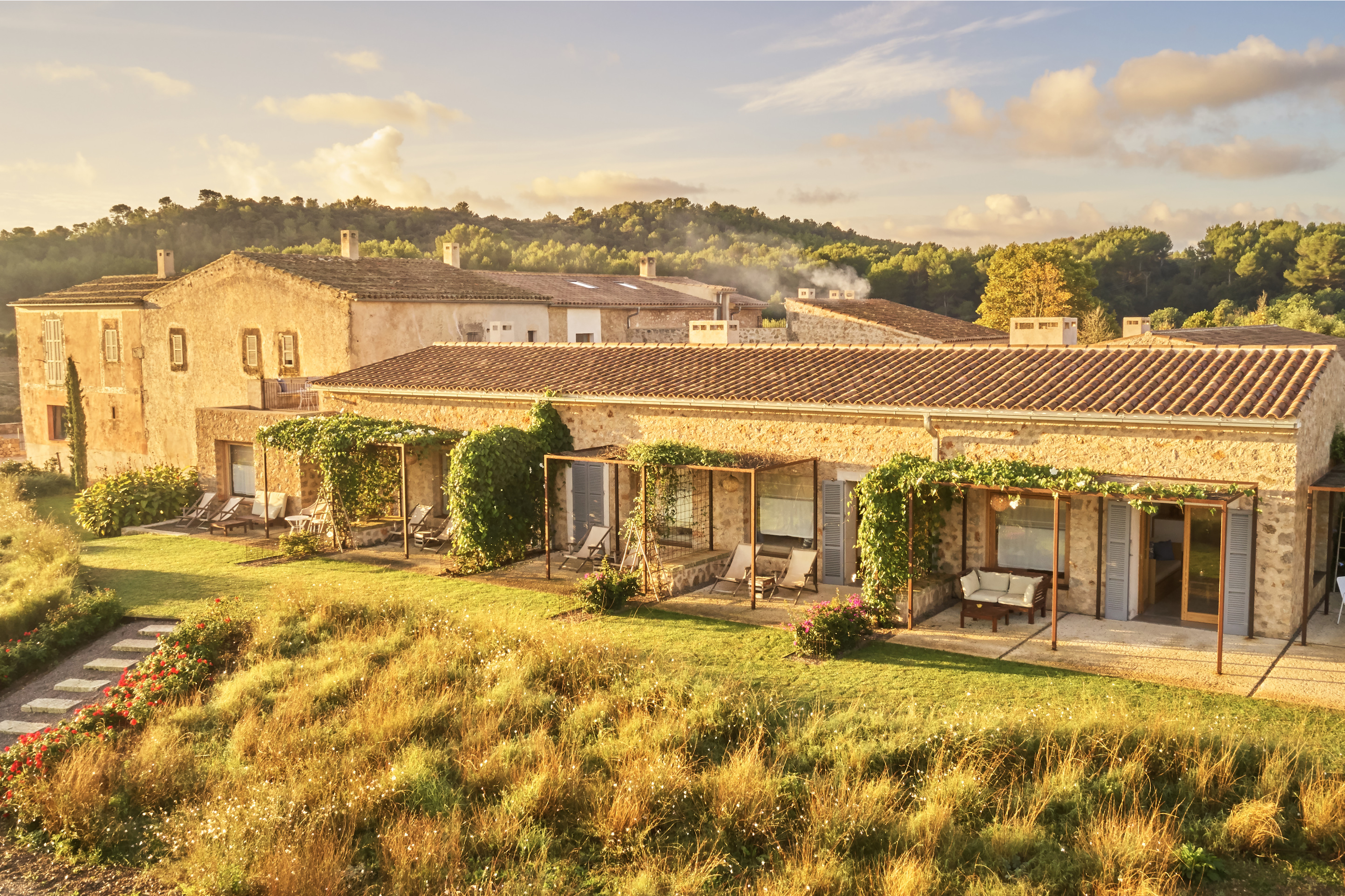 The long limestone facade of Finca Sestelrica seen from above at the end of the day, a regular run of shuttered bays and vine-covered pergolas above a green lawn, low wooded hills behind