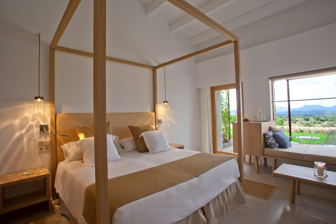 A bright finca room with a slim oak four-poster, white and stone-grey linen, and a wide window framing the hills and a sitting nook