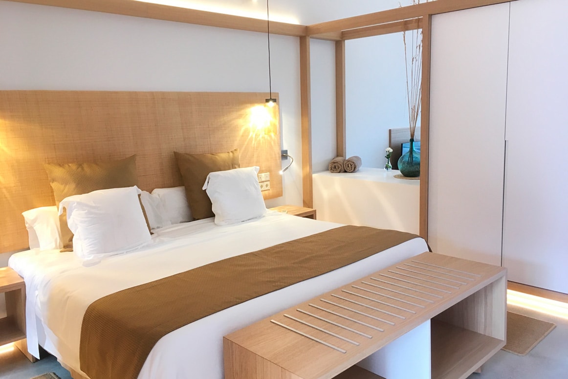 A warm-lit room at dusk with an oak headboard and bench, a tall mirror and a fitted oak wardrobe, lamplight raking across the plaster