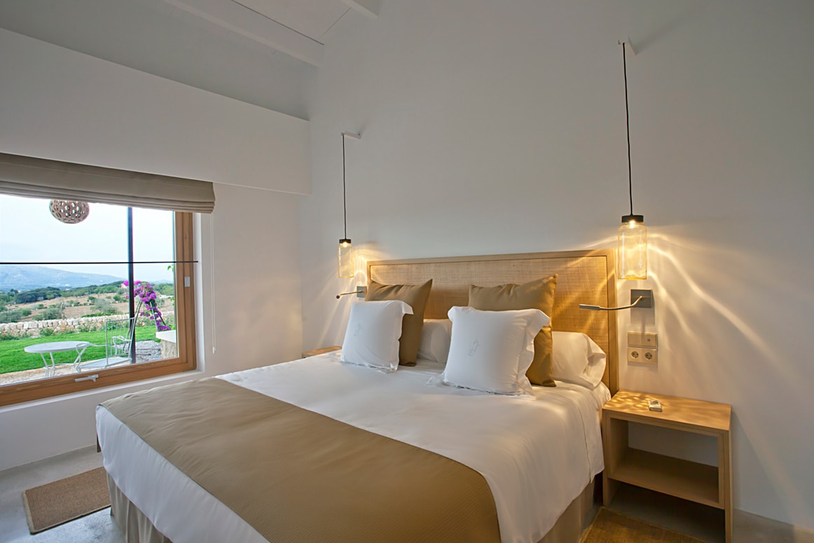 A calm double room with white walls, an oak headboard, two hanging glass lamps and a window onto the garden with bougainvillea and the hills