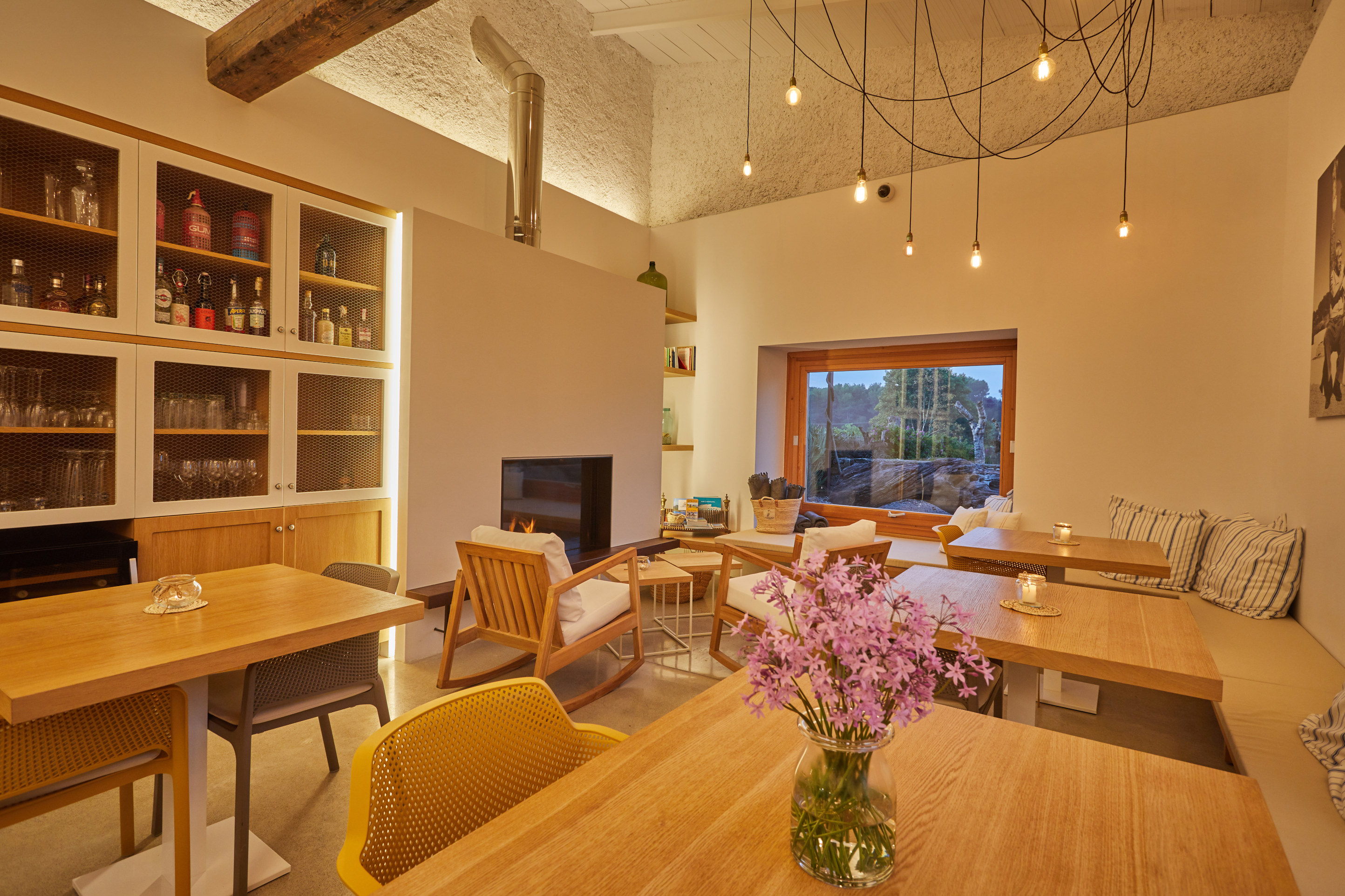 The finca dining room under a high beamed ceiling, a cluster of bare bulbs over oak tables, a fireplace and a wide window onto the garden