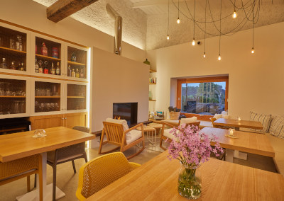 The beamed dining room with oak tables, a cluster of hanging bulbs and a fireplace, a window onto the garden