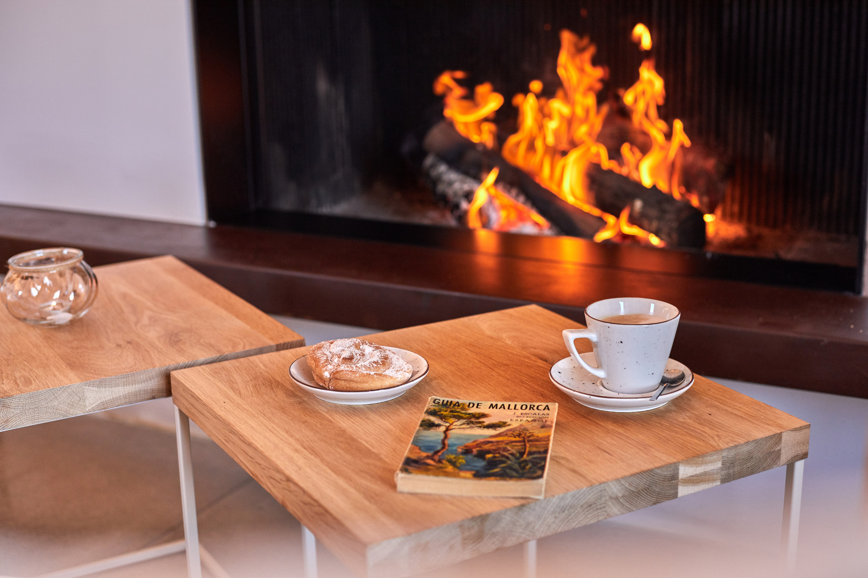 A wood fire behind glass with an oak table in front, an espresso and an old Guía de Mallorca laid on it