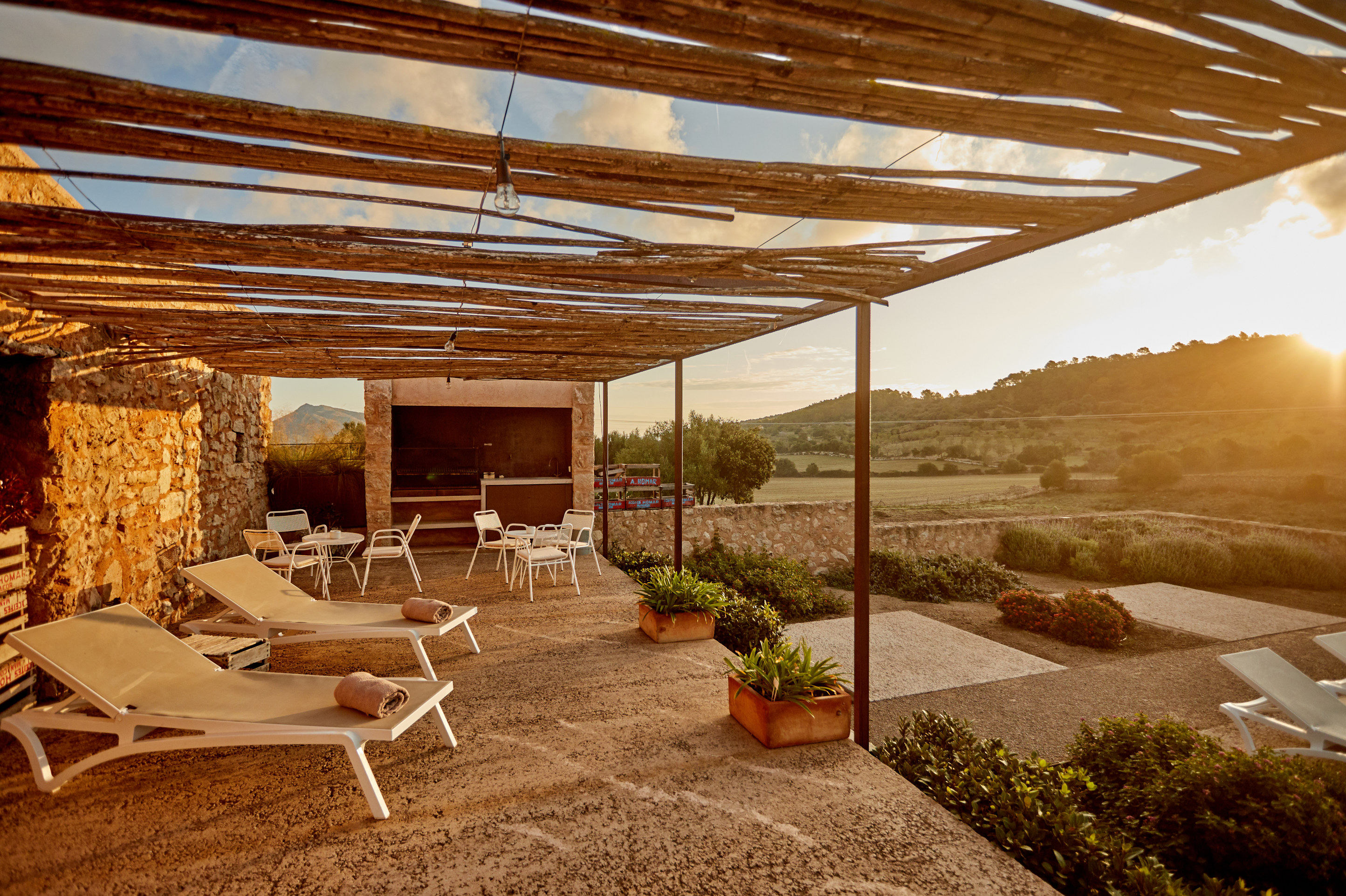 The courtyard terrace under a reed pergola at golden hour, loungers on stone paving, the open countryside beyond
