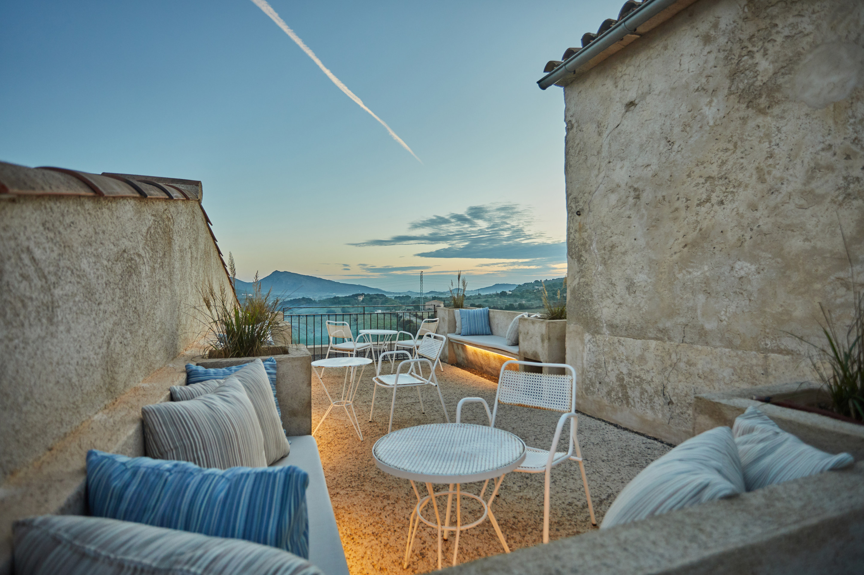 The stone roof terrace at blue hour, low cushioned benches and pale chairs between two limestone walls, looking out to the Tramuntana hills under a cool sky