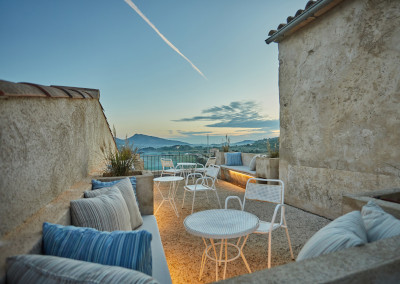 The roof terrace from the other side at dusk, pale metal chairs and a small table between stone walls, the hills going dark