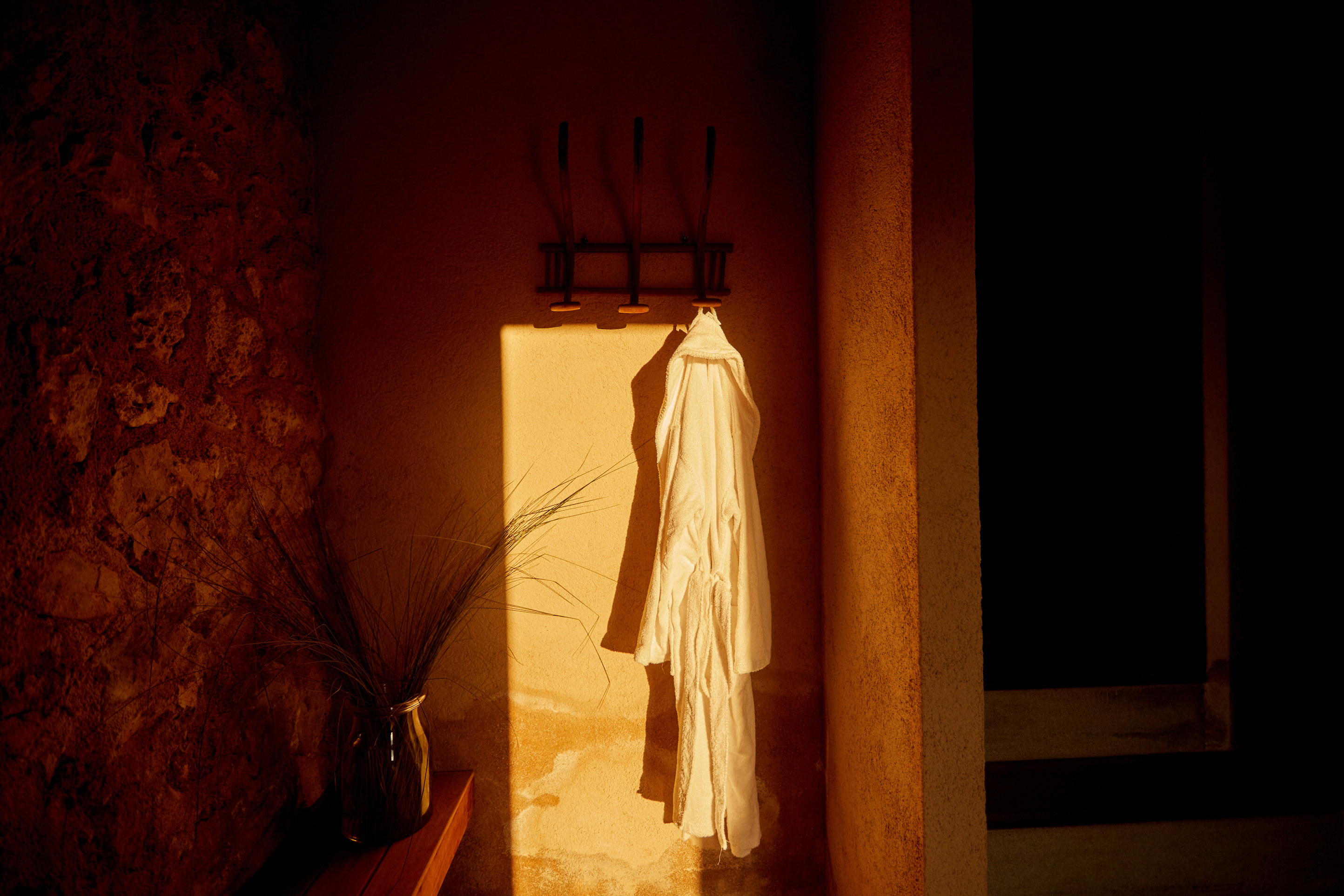 A shaft of low sun falling through a deep window onto lime plaster and rough limestone, a white robe and a bundle of dried grass caught in the light