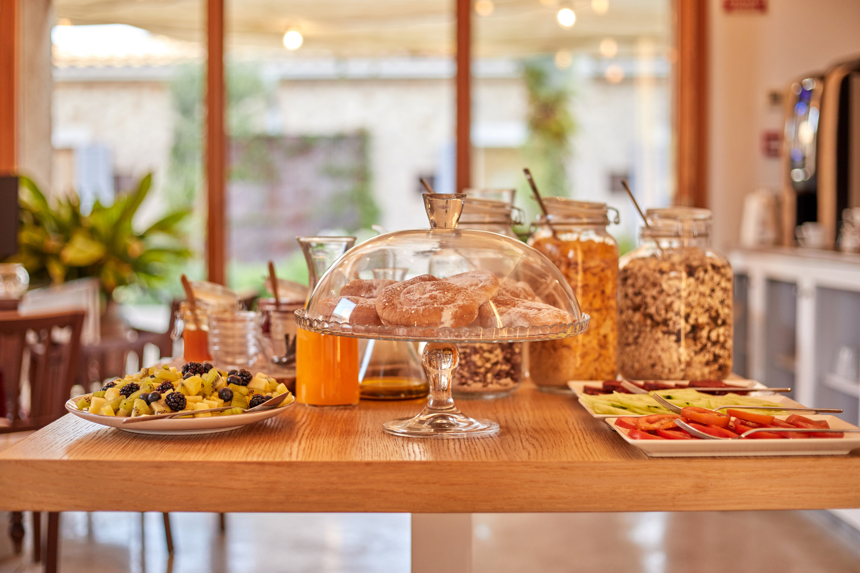 The breakfast table laid in the light: a glass cake stand of pastries, fruit, juice and jars of cereal against a wall of windows
